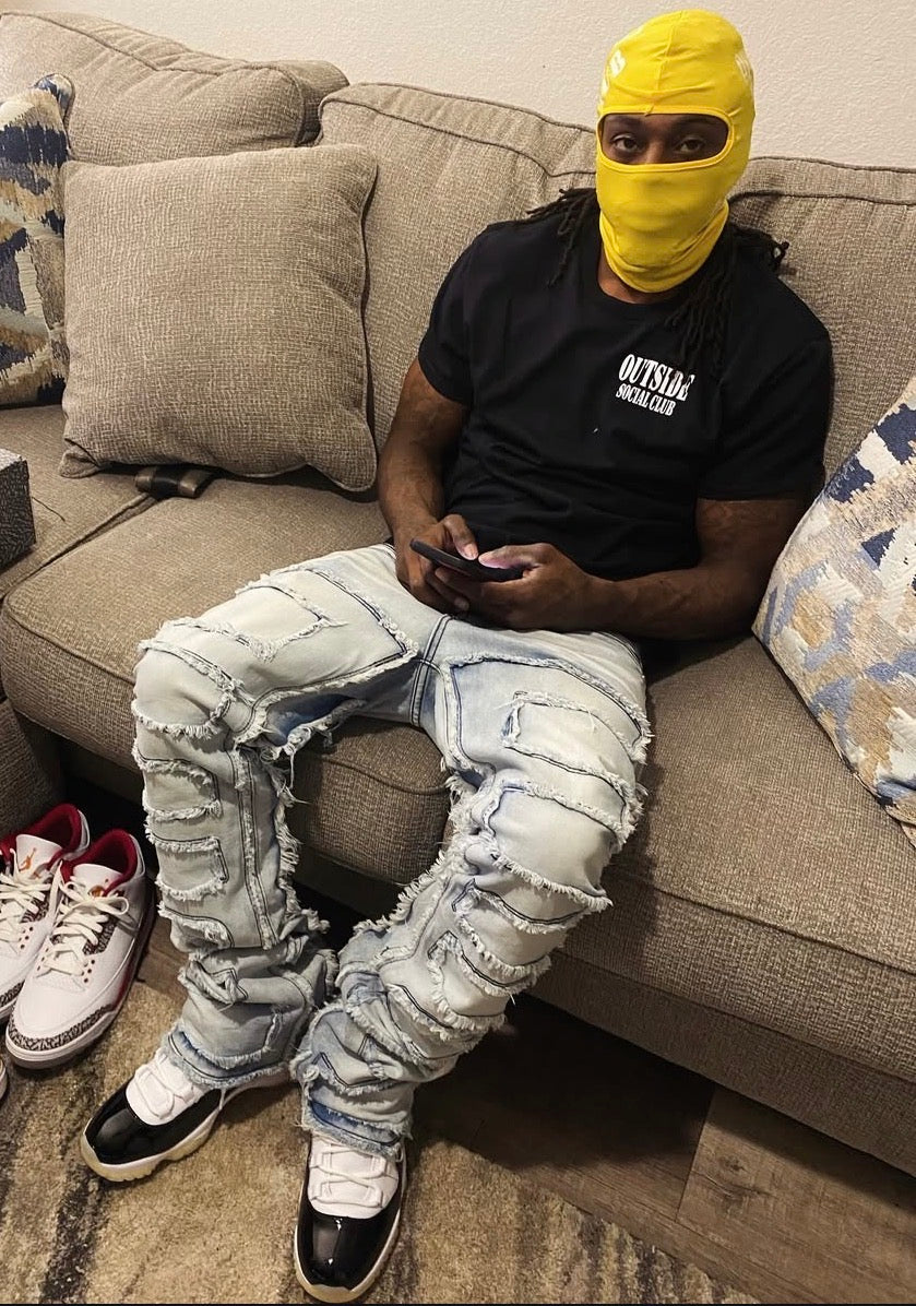 Person sitting on a couch wearing a yellow balaclava, black “outside socialclub “ t-shirt, ripped jeans, and sneakers.