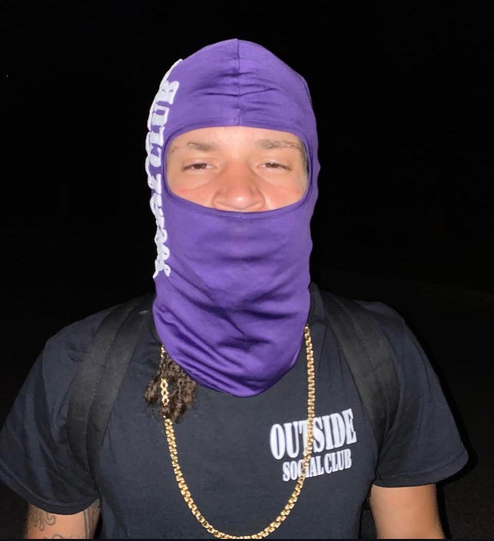 Person wearing a purple balaclava and black 'Outside Social Club' t-shirt on outside at night