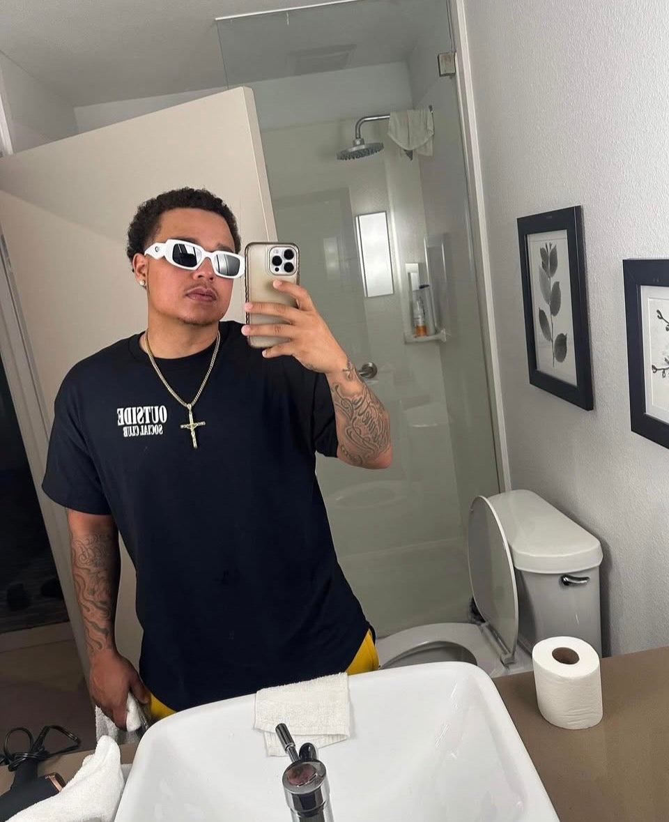 Person taking a mirror selfie in a bathroom wearing a black “outside social club” t-shirt with text and white sunglasses.
